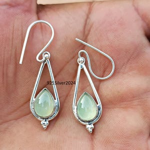 Puede incluir: Un par de pendientes de plata con dise&ntilde;o de l&aacute;grima. Cada pendiente presenta una piedra preciosa verde p&aacute;lido en forma de pera engastada en un marco de plata. Los pendientes tienen cierre de gancho y un peque&ntilde;o elemento decorativo. El texto "925Silver2024" es visible.