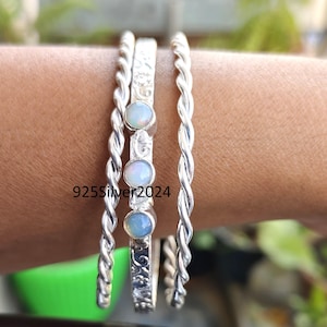 Può includere: Tre braccialetti d'argento. Due sono a forma di corda intrecciata, e uno è una fascia piatta con incisioni floreali e tre pietre preziose di opale. Il testo "925Silver2024" è visibile sulla fascia piatta.