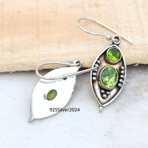 May include: A pair of sterling silver earrings with a leaf-shaped design. Each earring features two oval-cut, light green peridot gemstones. The earrings have a hook closure and are displayed on a wooden surface. The text "925Silver2024" is visible.