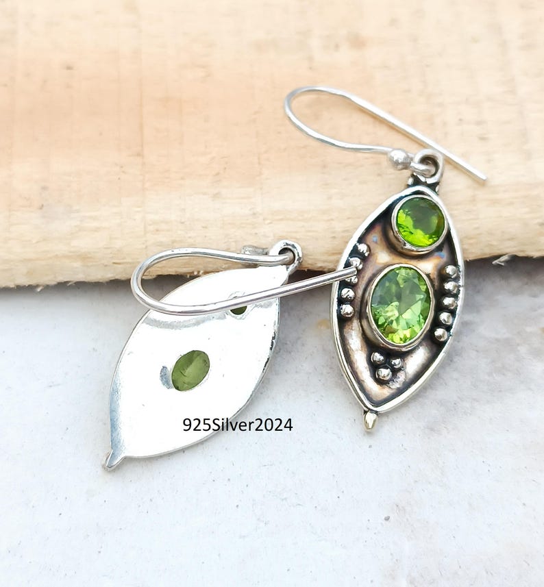 May include: A pair of sterling silver earrings with a leaf-shaped design. Each earring features two oval-cut, light green peridot gemstones. The earrings have a hook closure and are displayed on a wooden surface. The text "925Silver2024" is visible.