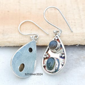 May include: Silver teardrop earrings with two oval-shaped gemstones. The earrings have a hook closure and are set against a light wooden background. The text "925Silver2024" is visible on one earring.
