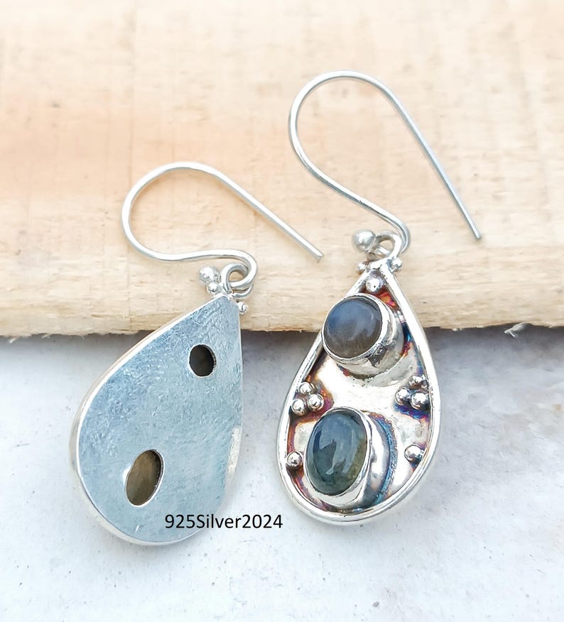 May include: Silver teardrop earrings with two oval-shaped gemstones. The earrings have a hook closure and are set against a light wooden background. The text "925Silver2024" is visible on one earring.