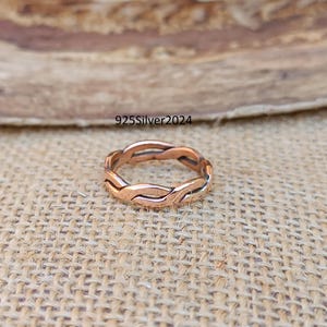 Puede incluir: Un anillo trenzado de oro rosa con un diseño sencillo. El anillo está sobre un fondo de arpillera marrón.