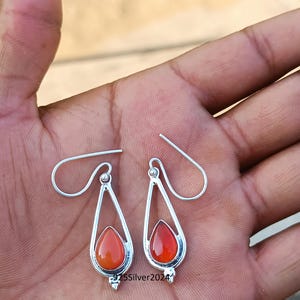 Puede incluir: Un par de pendientes plateados con forma de l&aacute;grima y piedras preciosas de color rojo anaranjado. Los pendientes tienen un dise&ntilde;o sencillo y elegante con cierre de gancho. El texto "925Silver2024" es visible.