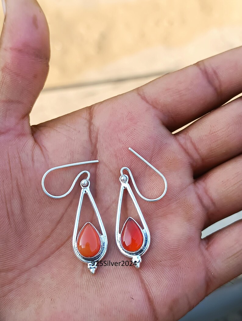 Puede incluir: Un par de pendientes plateados con forma de l&aacute;grima y piedras preciosas de color rojo anaranjado. Los pendientes tienen un dise&ntilde;o sencillo y elegante con cierre de gancho. El texto "925Silver2024" es visible.