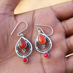 K&ouml;nnte beinhalten: Silberfarbene Ohrh&auml;nger mit roten, tropfenf&ouml;rmigen Steinen. Die Ohrringe haben ein detailliertes Design mit einem kleineren roten Stein unten und einem Hakenverschluss. Die Ohrringe werden auf einer Hand pr&auml;sentiert.