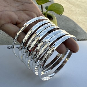 May include: A collection of silver bangles with a hammered finish. The bangles are circular and polished, reflecting light. The text "925Silver202" is visible on one of the bangles. The bangles are held in a hand.