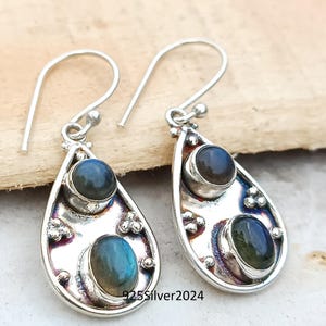 May include: A pair of silver teardrop earrings with two oval-shaped, blue-green gemstones. The earrings have a hook closure and are stamped with "925Silver2024". The gemstones have a shimmering, iridescent quality.
