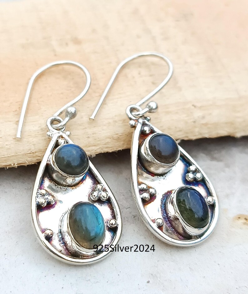 May include: A pair of silver teardrop earrings with two oval-shaped, blue-green gemstones. The earrings have a hook closure and are stamped with "925Silver2024". The gemstones have a shimmering, iridescent quality.