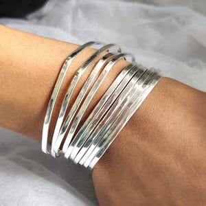 May include: A stack of silver bangles on a wrist. The bangles are thin, flat, and have a hammered texture. The bracelets are stacked together, creating a layered effect. The silver bangles are a simple and elegant accessory.