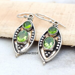 May include: Silver earrings with oval and round green gemstones. The earrings have a leaf-like shape with a beaded border and a hook closure. The text "925Silver2024" is visible on the earrings.