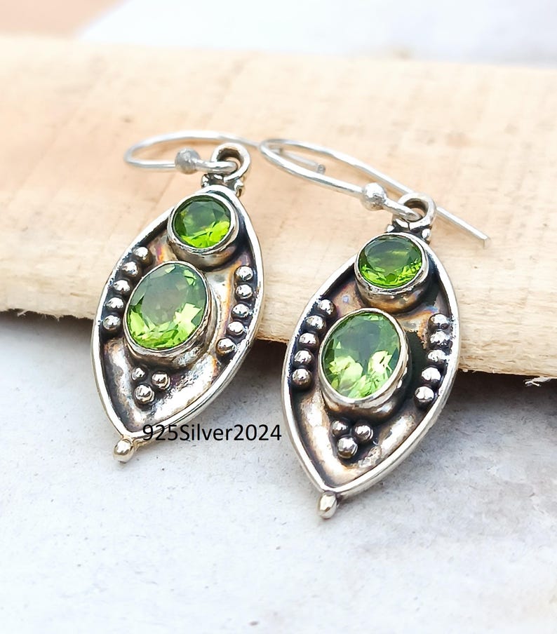 May include: Silver earrings with oval and round green gemstones. The earrings have a leaf-like shape with a beaded border and a hook closure. The text "925Silver2024" is visible on the earrings.