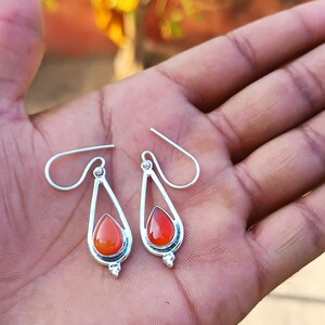 Puede incluir: Un par de pendientes plateados con forma de l&aacute;grima y una vibrante piedra preciosa de color rojo anaranjado en el centro. Los pendientes tienen un dise&ntilde;o sencillo y elegante con cierre de gancho. Las piedras son en forma de l&aacute;grima.