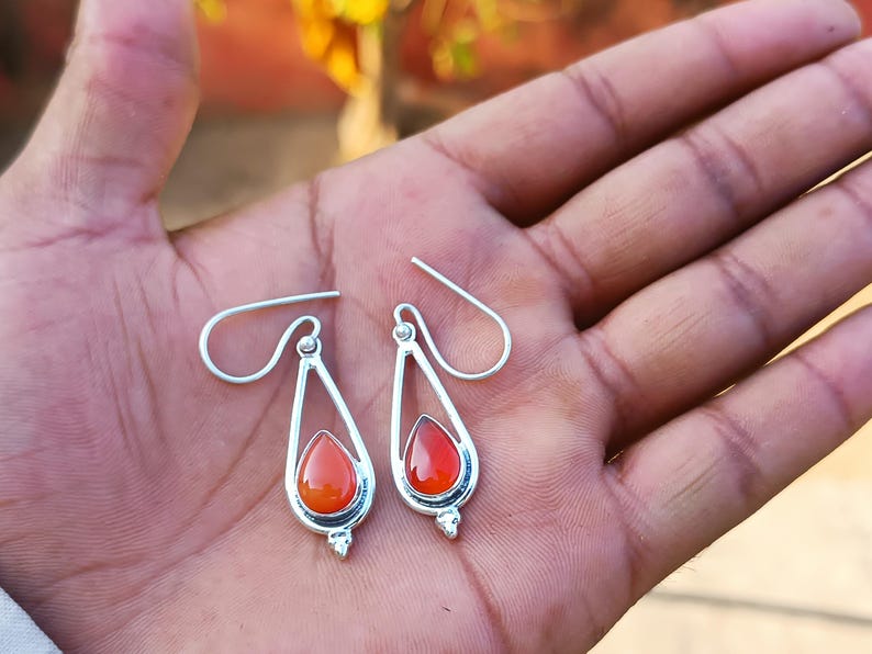 Puede incluir: Un par de pendientes plateados con forma de l&aacute;grima y una vibrante piedra preciosa de color rojo anaranjado en el centro. Los pendientes tienen un dise&ntilde;o sencillo y elegante con cierre de gancho. Las piedras son en forma de l&aacute;grima.