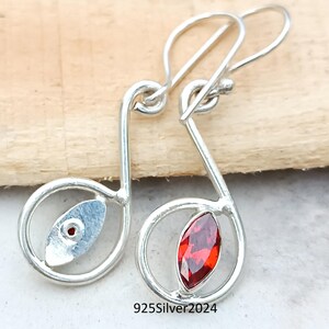 May include: A pair of silver earrings in the shape of musical notes. Each earring features a red gemstone and a small silver accent. The earrings are displayed on a light wooden surface. The text "925Silver2024" is visible.