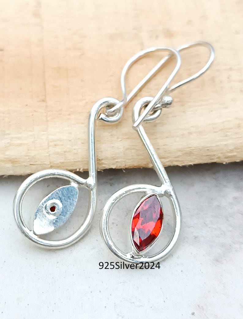 May include: A pair of silver earrings in the shape of musical notes. Each earring features a red gemstone and a small silver accent. The earrings are displayed on a light wooden surface. The text "925Silver2024" is visible.