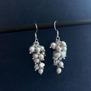 May include: A pair of dangle earrings featuring clusters of small, irregular, light gray freshwater pearls suspended from silver-toned metal hooks. Each earring consists of multiple pearls attached to silver chains, creating a cascading effect.