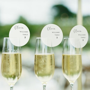 May include: Three champagne flutes filled with sparkling wine, each adorned with a white circular place card. The cards display the name "Olivia", the last name "Williams", and the table number "3". The background is blurred.