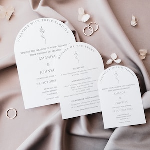 May include: Elegant wedding invitation suite featuring arched cards with floral accents and the names "Amanda & Johnson." The cards include event details and are set against a soft, neutral background with rings and floral decorations.