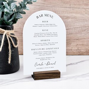 Puede incluir: Un menú de bar blanco arqueado con una base de madera oscura. El menú enumera cerveza, vino, licores y cócteles de autor. El texto incluye nombres de bebidas y la fecha de la boda.