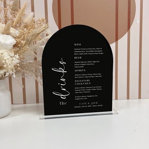 May include: Black arched wedding drink menu with white script text. The menu lists wine, beer, spirits, and signature cocktails. The menu includes the names of the drinks and the names of the couple, Cate & Jack, and the date and location of the wedding.