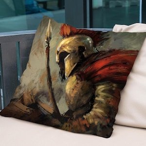 以下が含まれることがあります： 金色の兜と赤いマントを着た戦士が槍を持っている絵が描かれた装飾的な枕。戦士は劇的なポーズで描かれており、背景は落ち着いた色合いです。