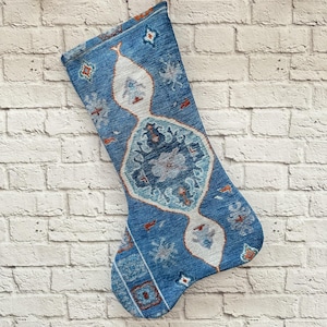 May include: A blue and white patterned Christmas stocking with a geometric design. The stocking is hanging from a wooden hook on a white brick wall.