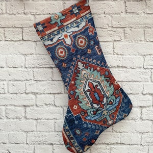 May include: A blue and red Christmas stocking with a traditional rug pattern. The stocking is hanging on a wooden hook against a white brick wall.