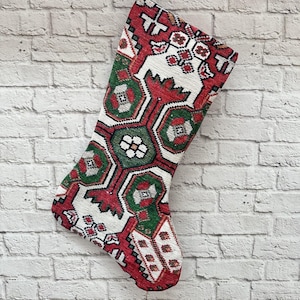 Puede incluir: Una media de Navidad roja, verde y blanca con un diseño tradicional. La media está colgada de un gancho de madera en una pared de ladrillo blanco.