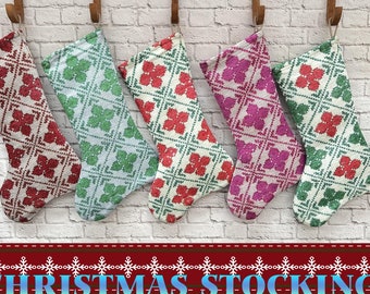 Puede incluir: Cinco calcetines de Navidad colgados de ganchos en una pared de ladrillo blanco. Los calcetines están hechos de tela blanca con un patrón de diamante y presentan acentos de diferentes colores: rojo, verde, rosa y azul.