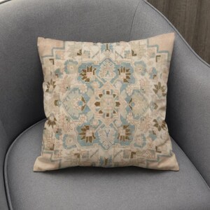 May include: A square throw pillow featuring a symmetrical, intricate pattern in shades of light blue, beige, and soft brown. The design consists of geometric and floral-inspired motifs arranged in a central medallion layout on a cream background.