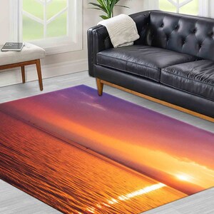 Op de afbeelding: Een rechthoekig vloerkleed met een zonsondergang boven de oceaan. Het ontwerp van het vloerkleed gaat over van diep oranje naar paars, met de weerspiegeling van de zon op het water. Het vloerkleed ligt op een lichtgekleurde houten vloer.