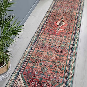 May include: A long, rectangular rug with a distressed, vintage look. The rug features a central design in shades of red, cream, and teal, with a detailed border. The rug is placed on a light-colored wooden floor.