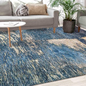 May include: A blue and cream abstract area rug with a contemporary design. The rug is laid out on a hardwood floor in a living room setting with a white sofa and coffee table.