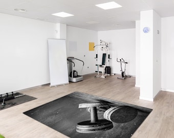 May include: Black and white image of a home gym with exercise equipment. A dumbbell is in the foreground on a black and gray rug. Other equipment includes a weight machine, exercise bike, and a vibration plate.