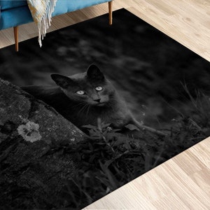 May include: A black cat with bright green eyes sits in a dark, shadowy forest setting. The cat is looking directly at the camera, with its head tilted slightly to the side. The background is a blur of dark green and brown foliage.