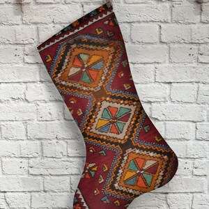 Puede incluir: Un calcetín navideño con un diseño de alfombra tradicional. El calcetín presenta un fondo rojo oscuro con patrones geométricos en tonos naranja, amarillo, azul y verde. Cuelga de un gancho de madera contra una pared de ladrillos blancos.