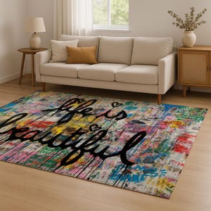 May include: A rectangular area rug with a colorful, abstract design. The rug features a mix of paint splatters and graffiti-style text that reads "Life is Beautiful" in black script. The rug is placed on a wooden floor.