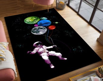 Puede incluir: Alfombra rectangular negra con un astronauta flotando con globos con forma de planetas de colores. La alfombra tiene un tema espacial con un fondo estrellado y un diseño caprichoso. El astronauta lleva un traje espacial blanco.