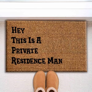 May include: A rectangular doormat made of natural fibers with the text "HEY THIS IS A PRIVATE RESIDENCE MAN" in black block letters. The mat is placed in front of a white door and a pair of brown slippers.