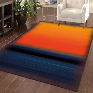 May include: A rectangular area rug with an abstract design. The rug features a gradient of colors, transitioning from yellow to orange, then to a dark blue at the bottom, with a black horizontal stripe. The rug is placed on a wooden floor.