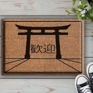 May include: A rectangular doormat with a brown, textured surface. A black graphic of a Japanese torii gate and Japanese characters are centered on the mat. Black and white trainers are next to the mat.