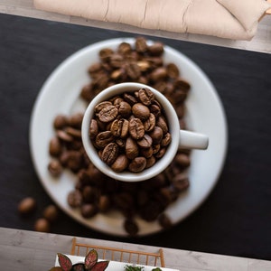 May include: A white ceramic cup filled with roasted coffee beans sits on a white saucer surrounded by more coffee beans. The cup and saucer are on a black surface.