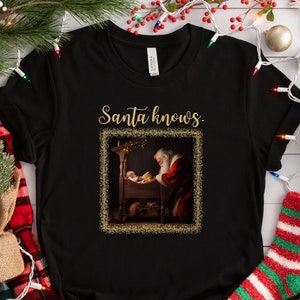 May include: Black t-shirt with a gold glitter design featuring a picture of Santa Claus kneeling beside a baby in a manger. The text "Santa knows." is above the picture.