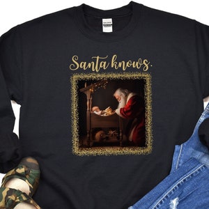 May include: Black sweatshirt with the text "Santa knows." in gold script. The design features an image of Santa Claus kneeling beside a baby in a manger, framed with a gold glitter border. The sweatshirt is a Gildan brand.