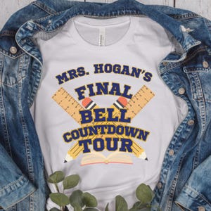 May include: White t-shirt with a blue and yellow graphic design that says "Mrs. Hogan's Final Bell Countdown Tour". The graphic features two pencils crossed over each other with rulers and a book.