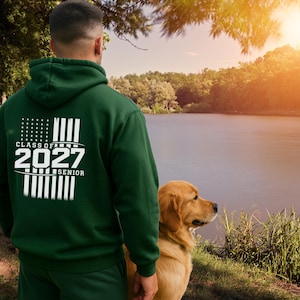 May include: A dark green hooded sweatshirt featuring a white graphic on the back. The design includes a stylised American flag pattern with the text "CLASS OF 2027 SENIOR" printed in bold white lettering across the centre of the flag graphic.