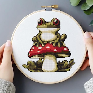 May include: Cross-stitch embroidery of a green frog with red eyes sitting on a red mushroom with white spots. The embroidery is in a wooden hoop, a craft project with a whimsical theme.