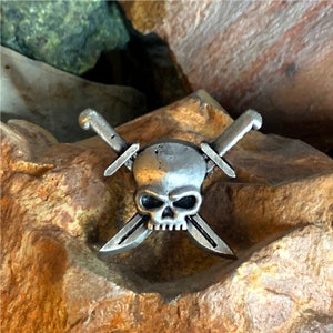 Puede incluir: Un pin de calavera de plata con dos espadas cruzadas. La calavera tiene un diseño detallado y las espadas son simples.
