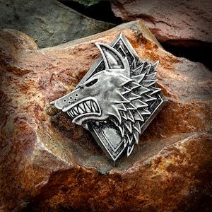 May include: A silver-toned metal pin featuring a detailed wolf head design. The wolf's mouth is open, showing sharp teeth, and its fur is textured. The pin is shaped like a diamond and has a dark grey background.